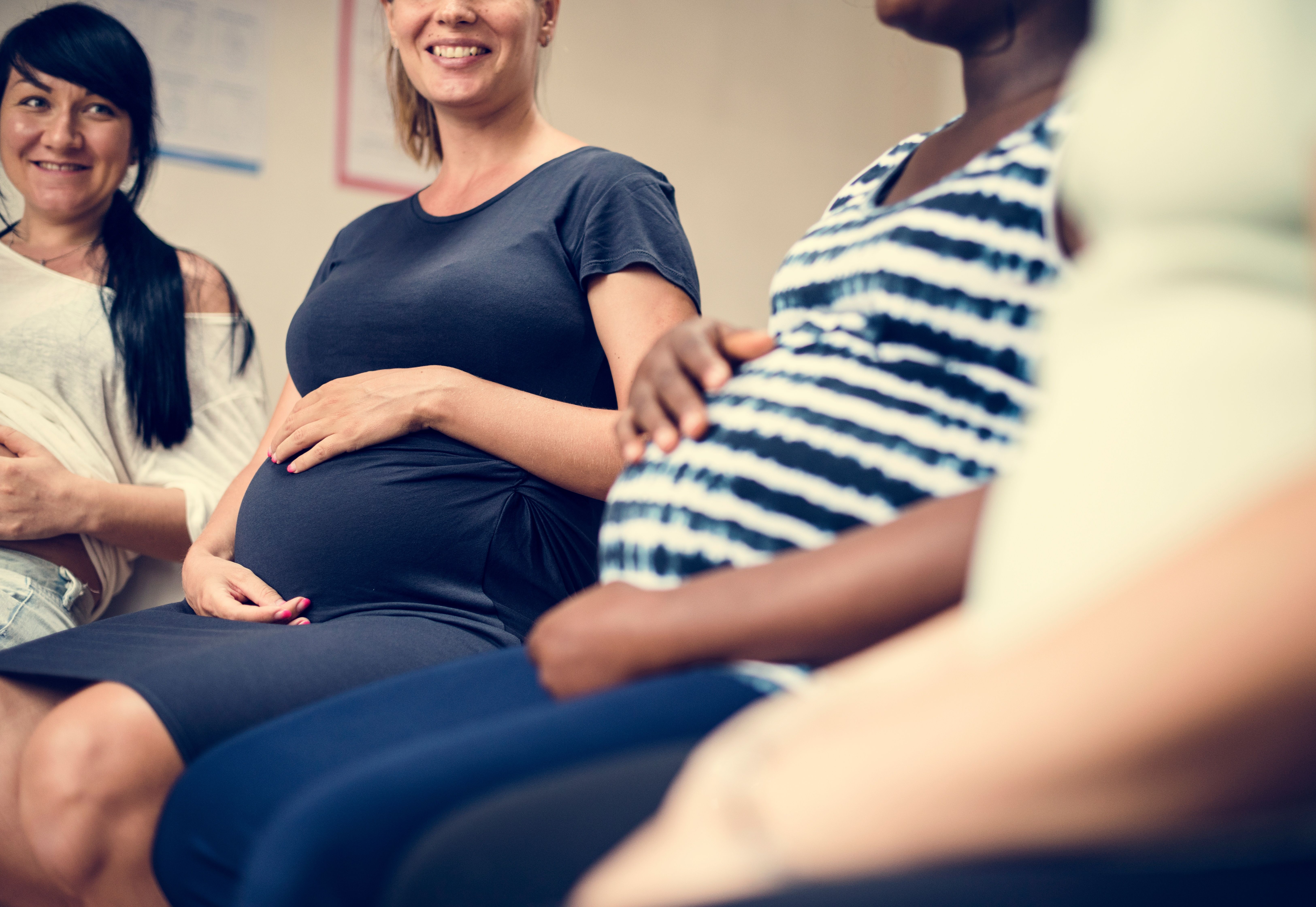 A group of pregnant women sit together. Two of them are seen smiling and all are holding their bellies.